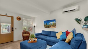 A living room with a blue sectional sofa, a small side table with a tray, a wicker chair, and a large window with blinds. - The Tucsonan Style Casita - FULLY FENCED AND PET FRIENDLY! (Tucson)