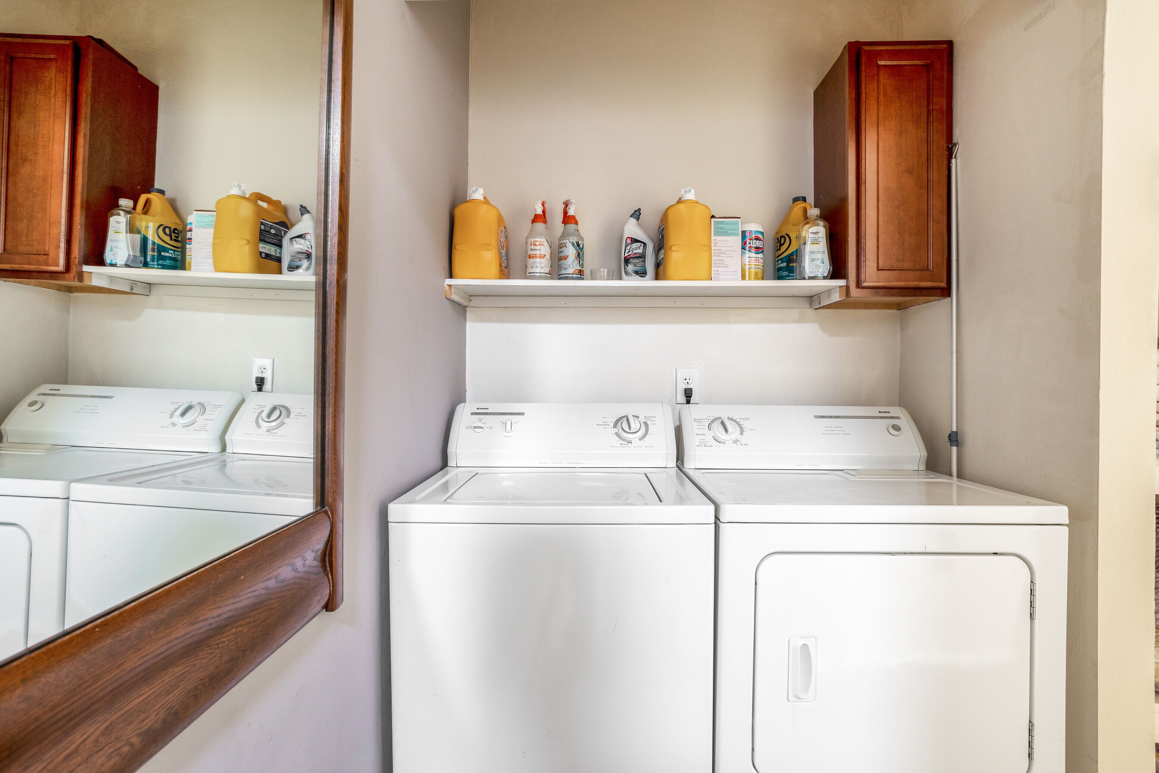 A laundry area with two white washers and dryers, wooden cabinets above, and shelves with cleaning supplies.