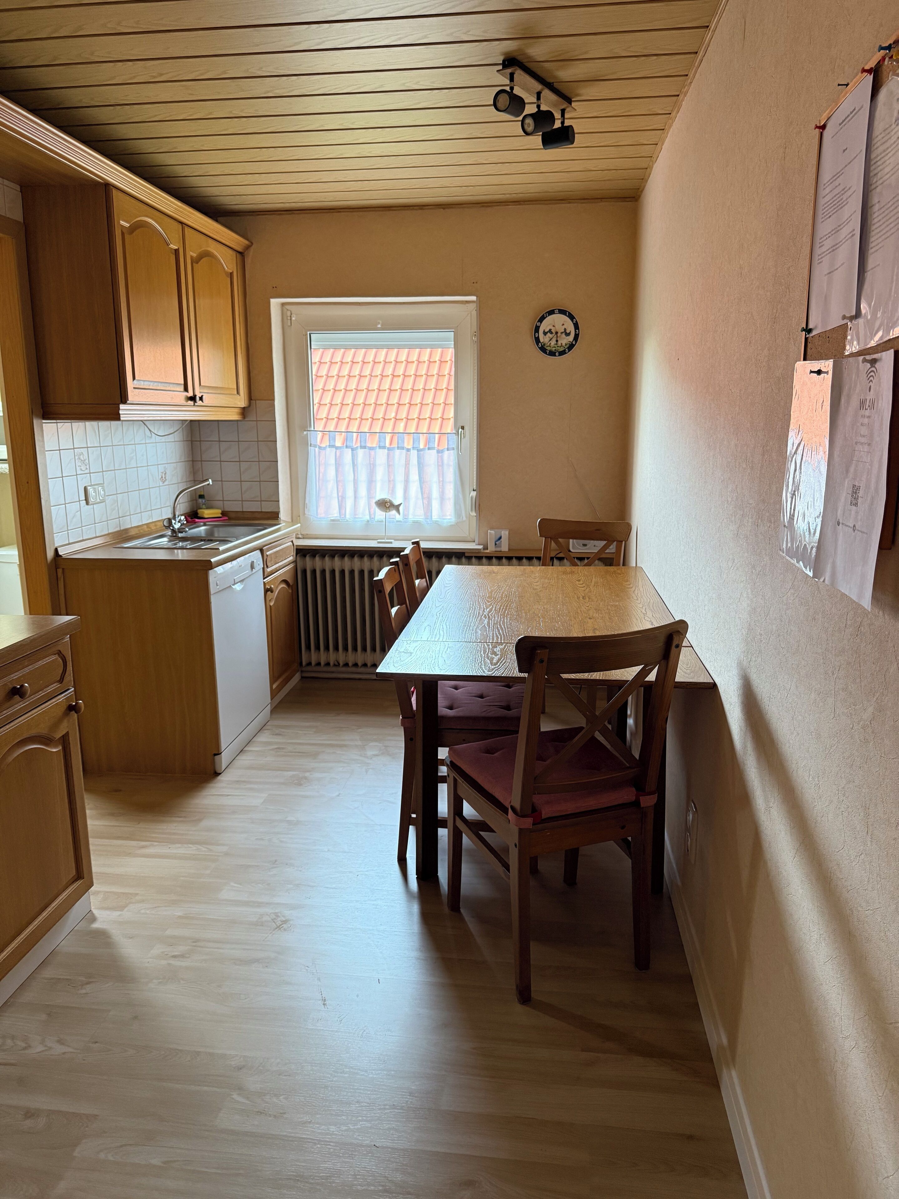 Ett kompakt kök med träskåp, en inbyggd diskmaskin och ett fönster med utsikt över ett rött tegeltak.