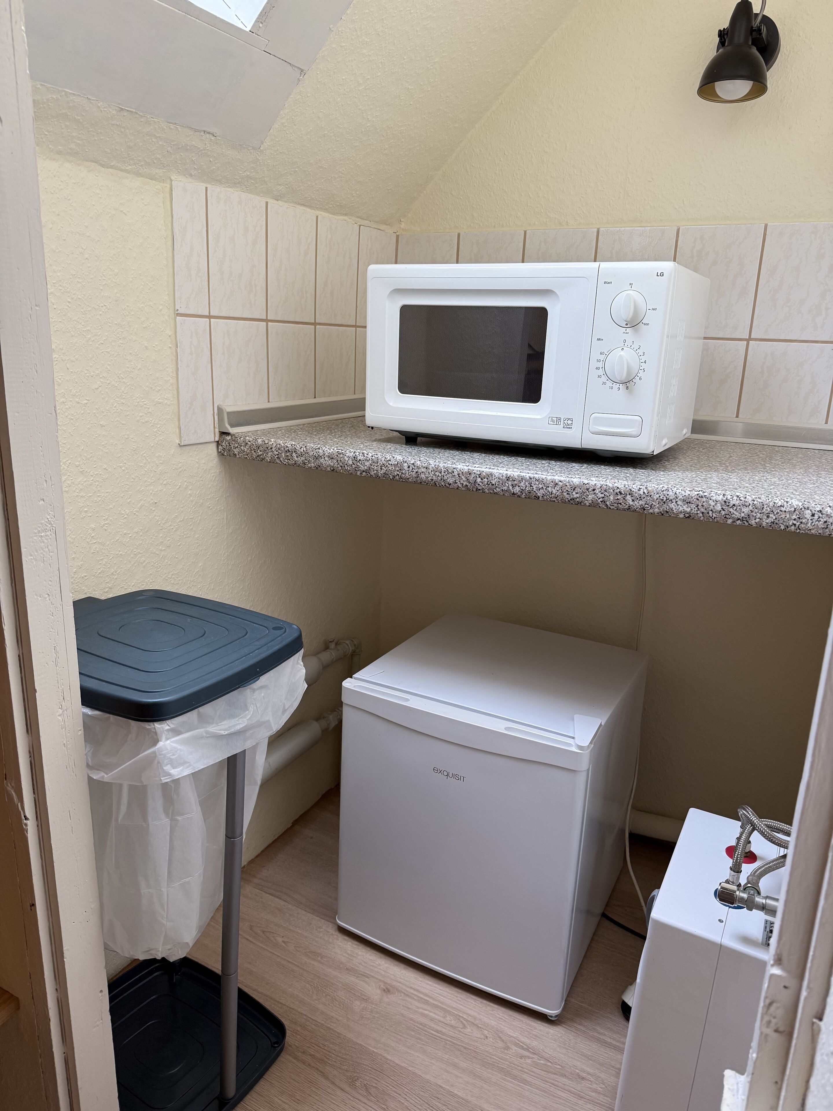 A compact kitchenette with a white microwave on a countertop, a small refrigerator below, and a trash bin with a lid.