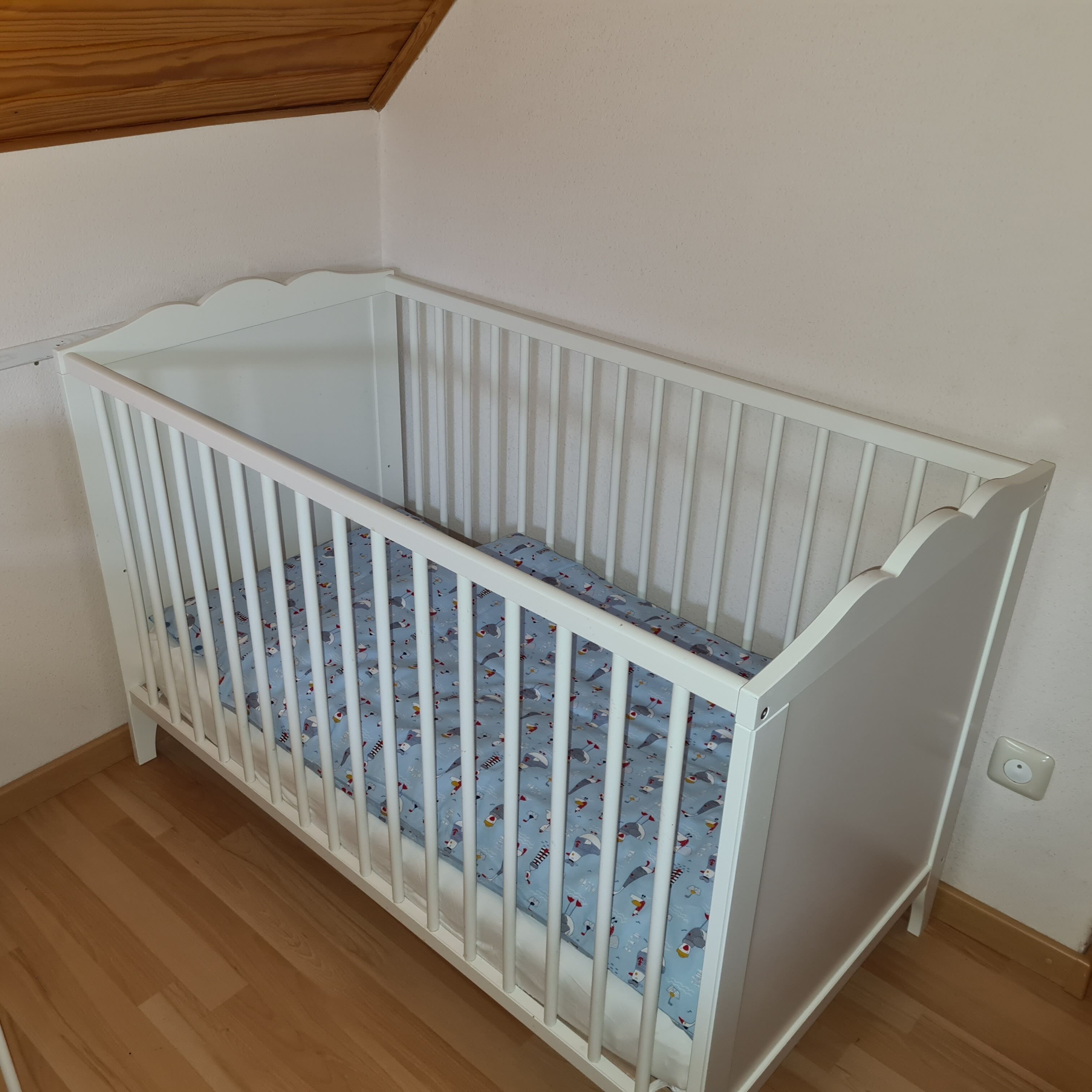 Un berceau blanc avec un matelas bleu orné de motifs d’animaux, placé dans un coin d’une pièce dont le sol est en bois et le plafond est en pente.