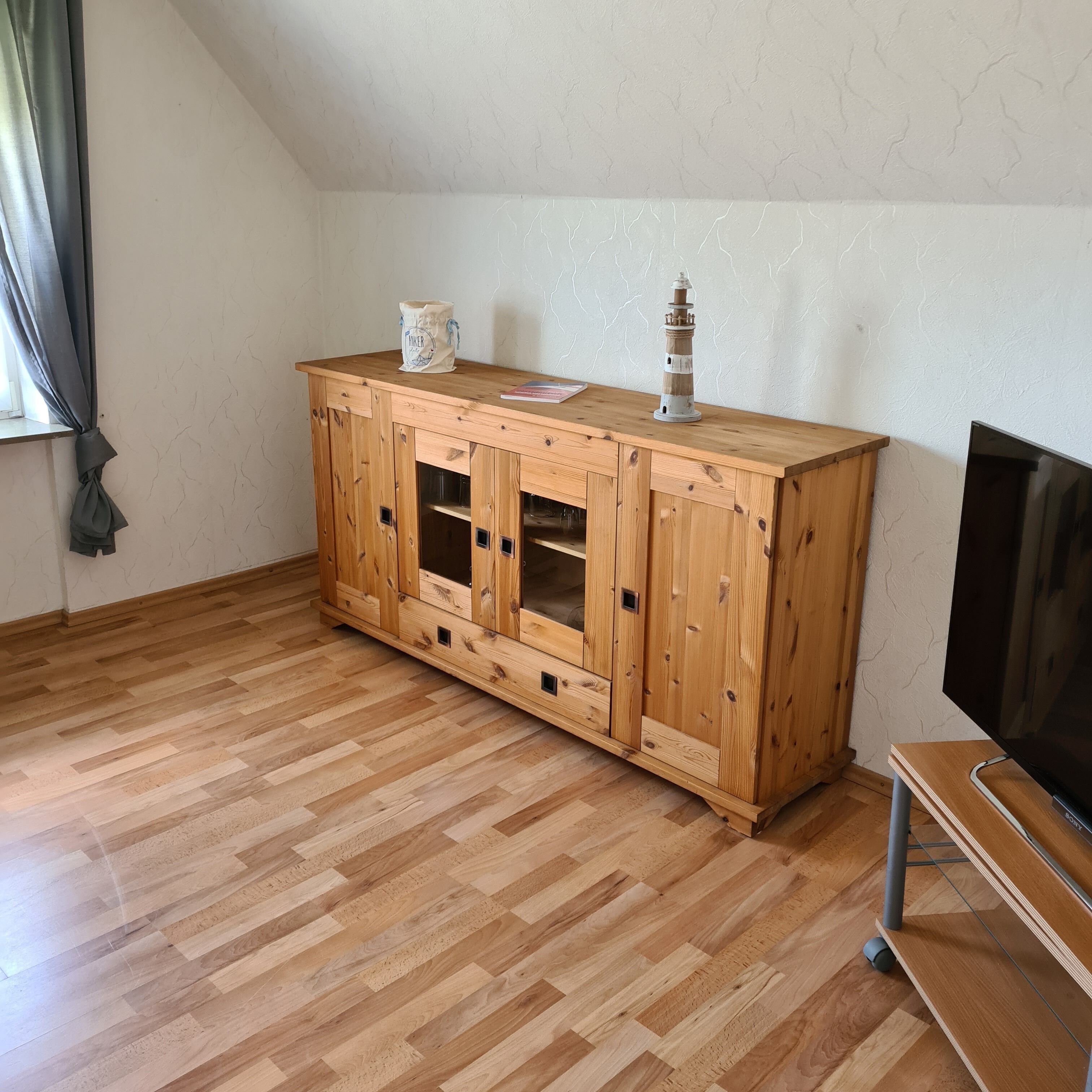 A wooden cabinet with glass doors, a small table with a lamp, and a TV stand in a room with wooden flooring and a sloped ceiling.