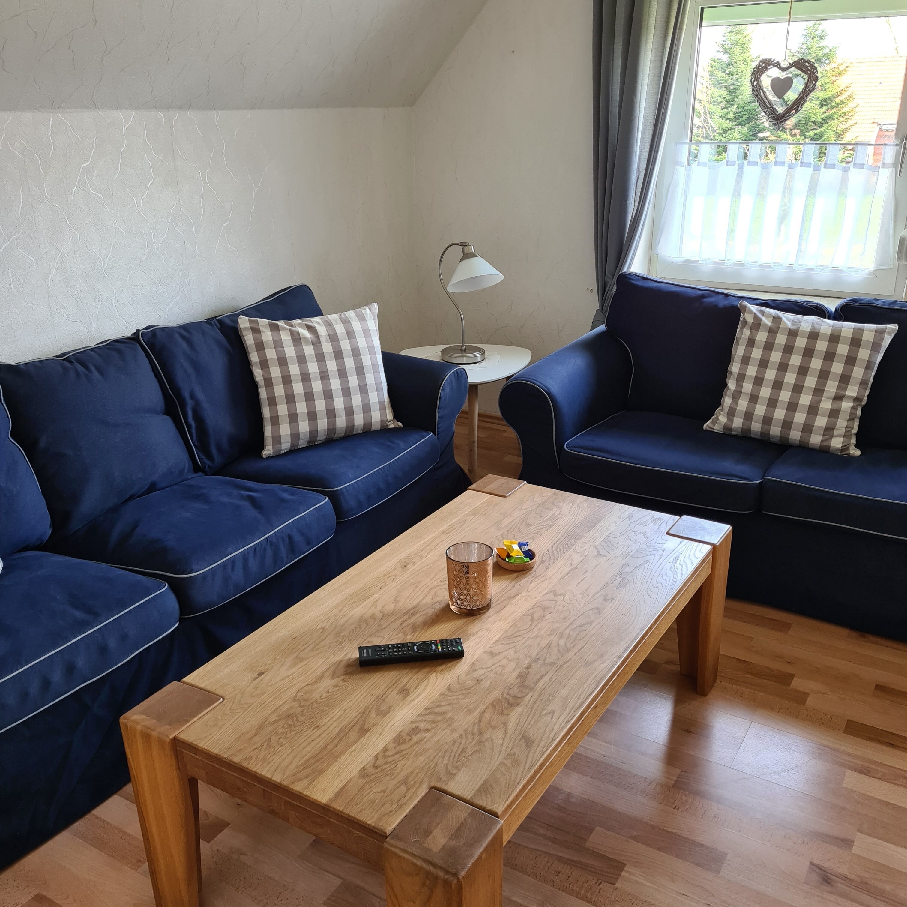 Een woonkamer met twee blauwe banken, een houten salontafel, een klein bijzettafeltje met een lamp, en een raam met een hartvormige decoratie.