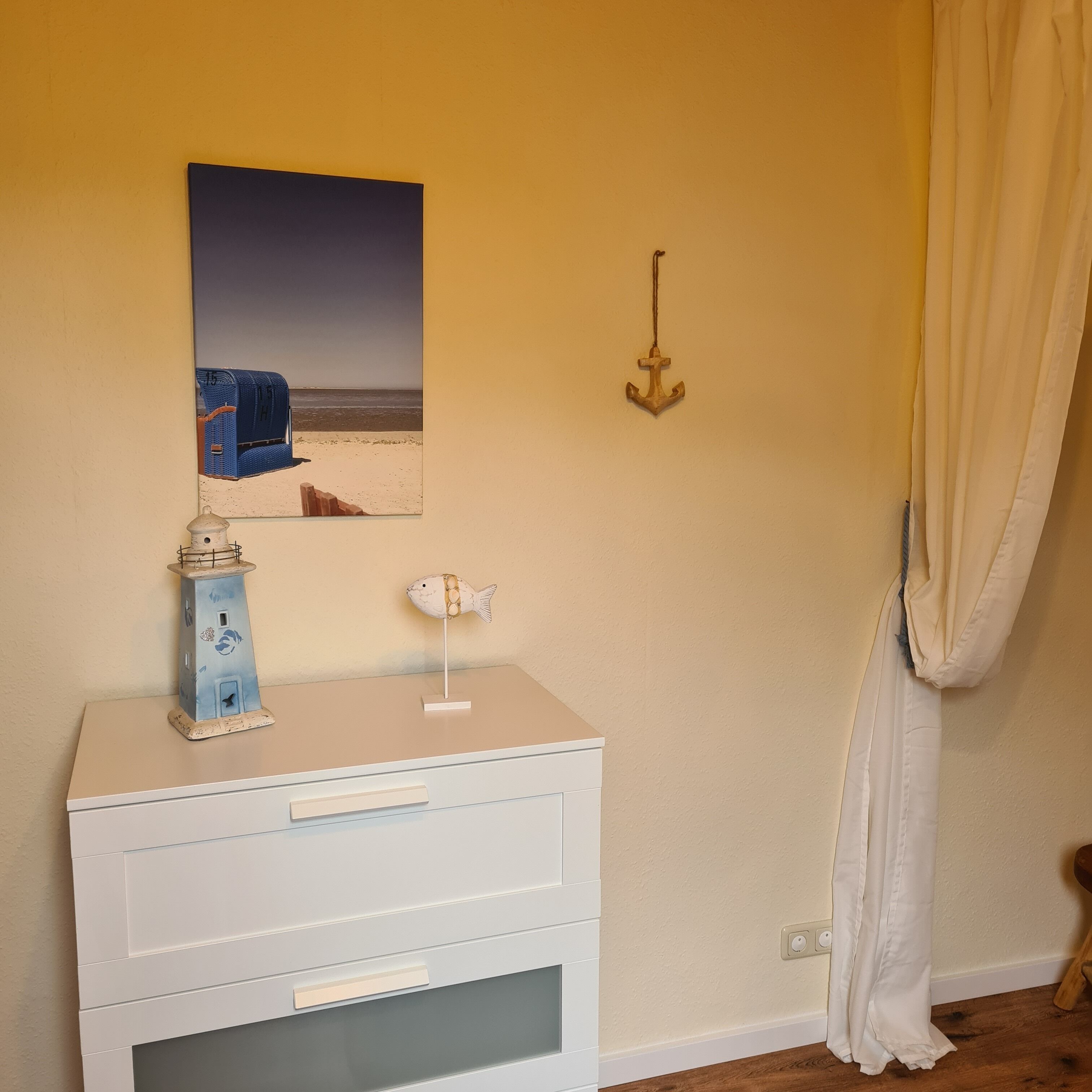 Une pièce comprenant une commode blanche, un objet bleu et blanc en forme de poisson, une petite maquette de phare et un tableau représentant une scène de plage.