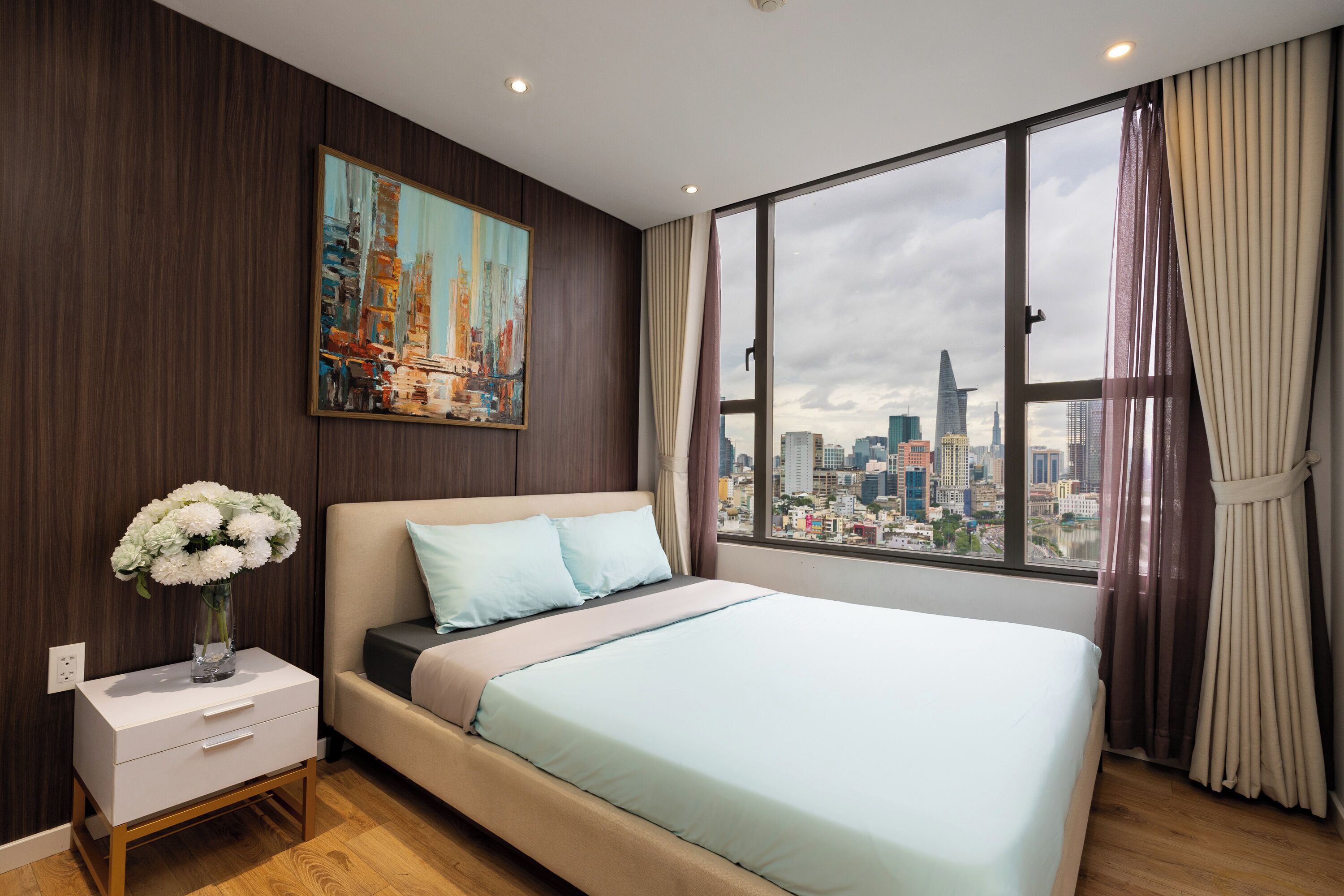 A bedroom with a bed, a nightstand with a vase of flowers, a painting on the wall, and a large window with a city view.