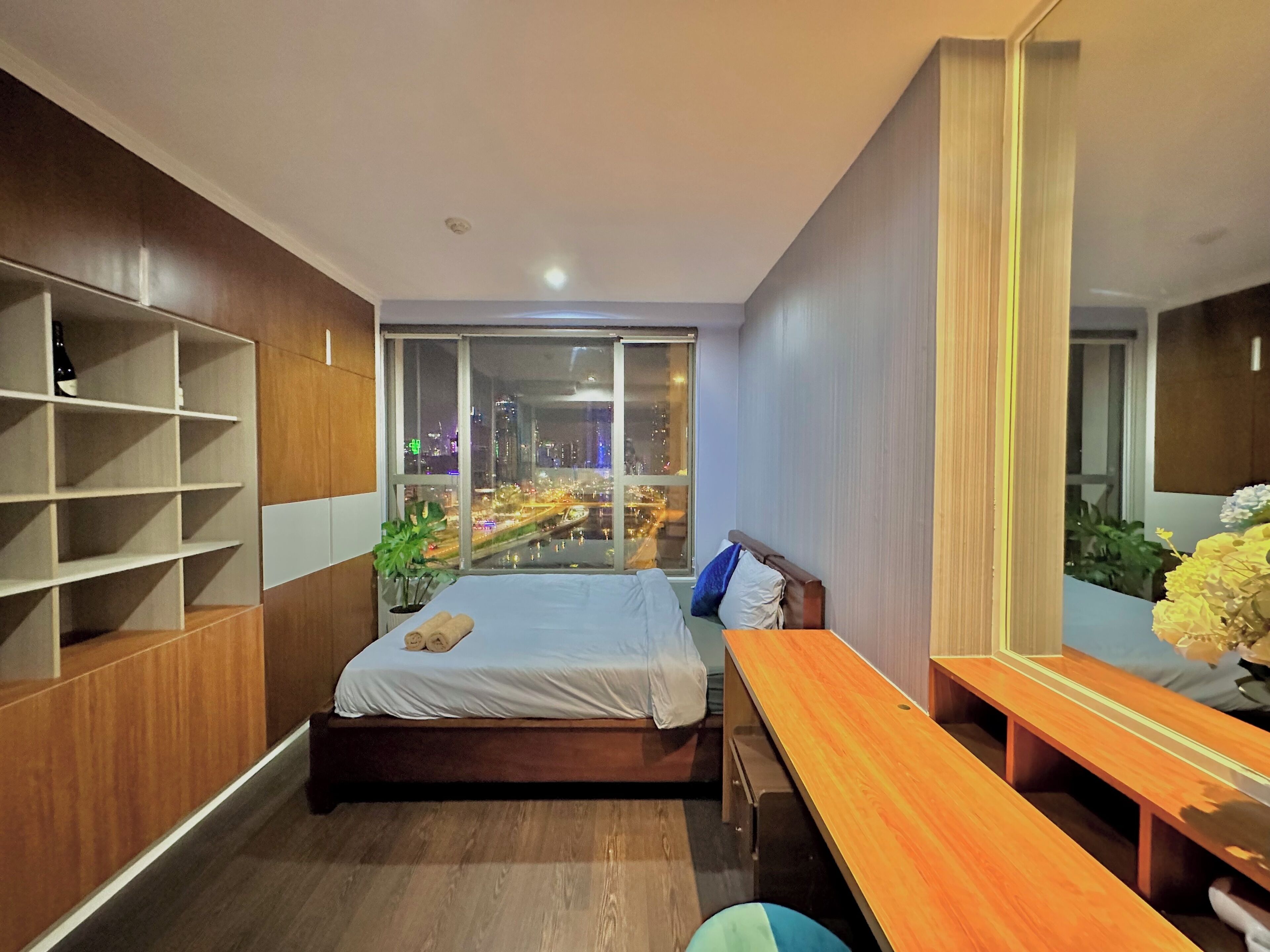 A modern bedroom with a bed, a wooden desk, a large mirror, and a city view through a window.