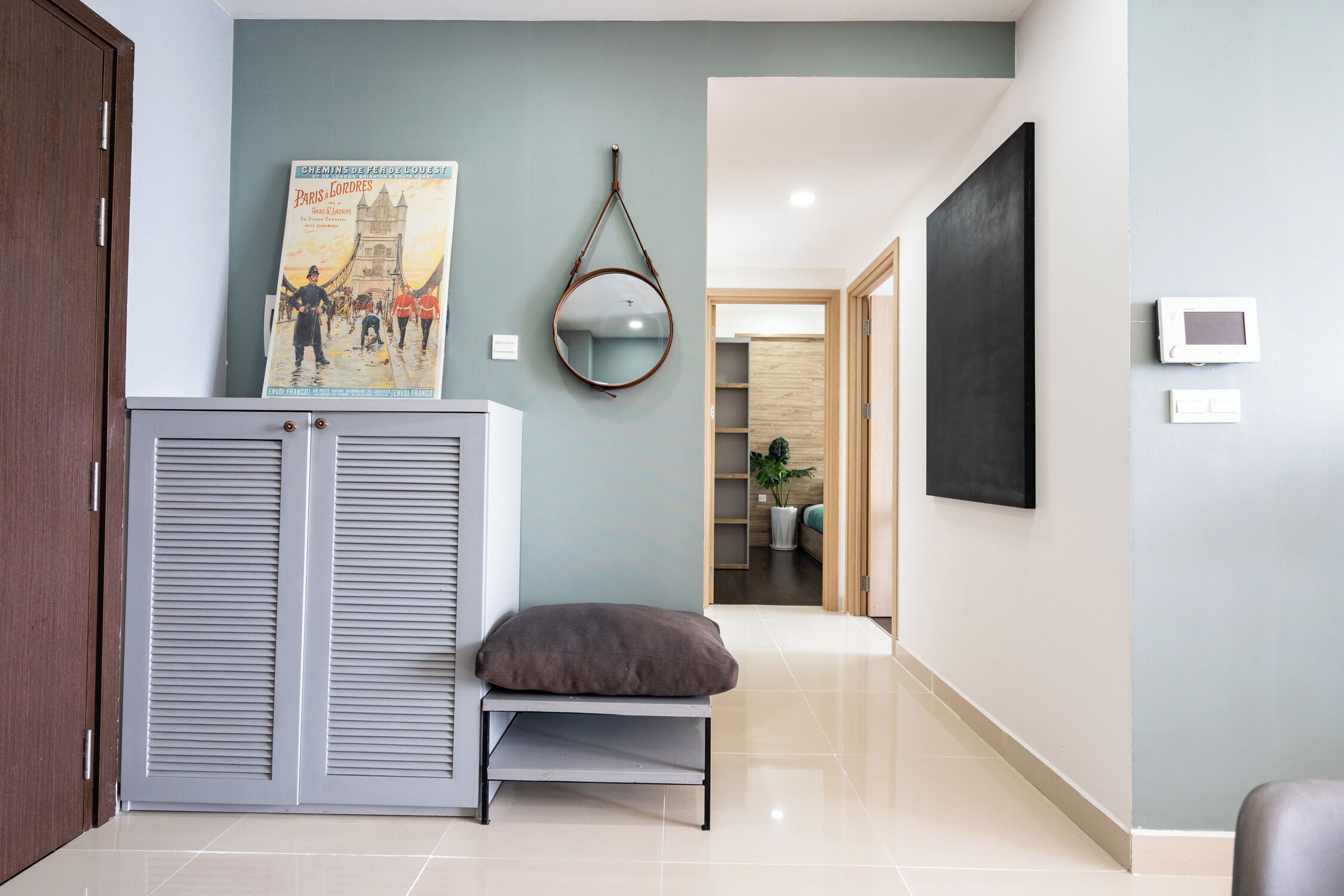 A hallway with a grey bench, a poster on the wall, a mirror, and a door leading to another room.
