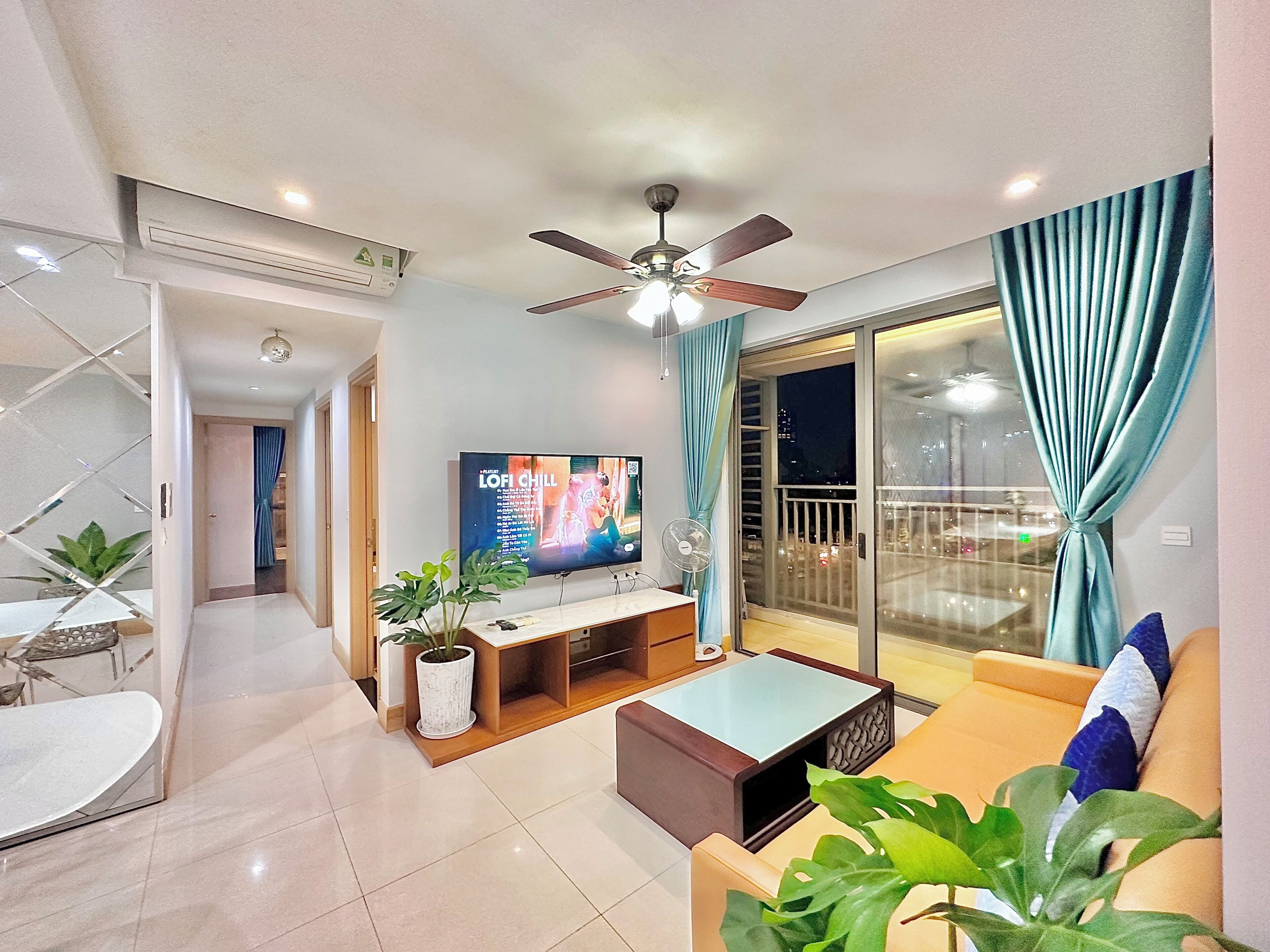 A modern living room with a flat-screen TV, a ceiling fan, a sliding glass door to a balcony, and a dining area with a table and chairs.