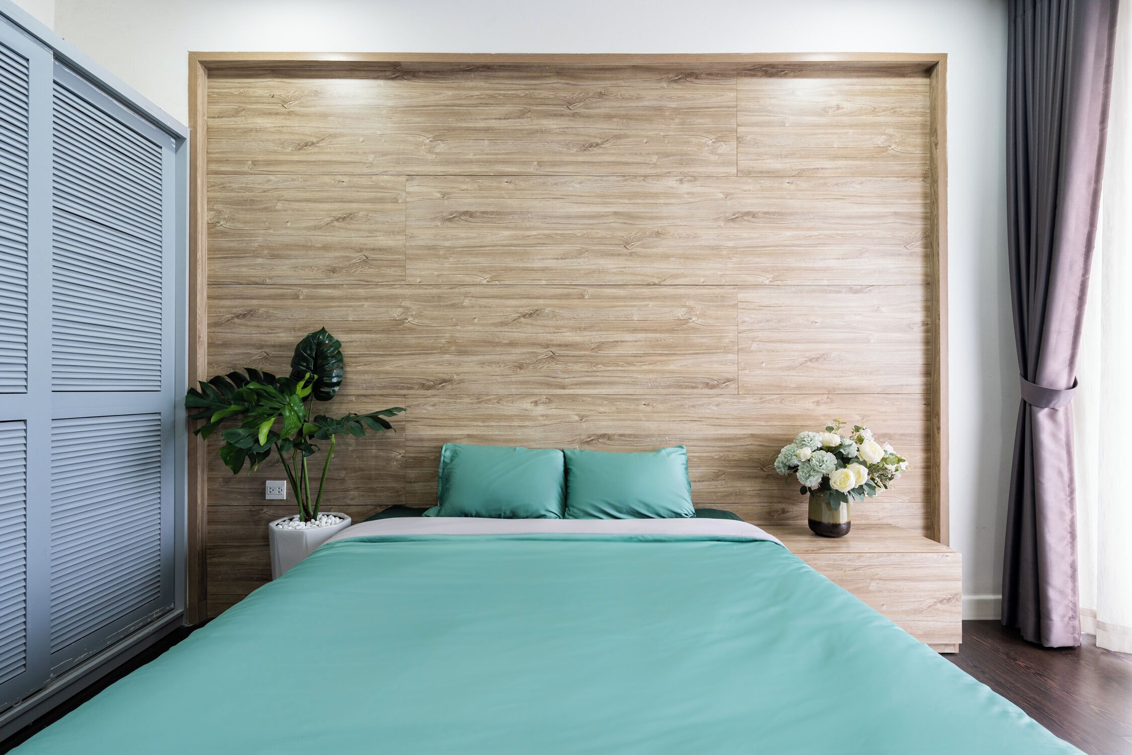 A bedroom with a wooden headboard, a bed with teal bedding, a potted plant, and a vase with flowers.