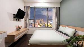 A hotel room with a bed, a TV mounted on the wall, a window with curtains, and a night view of the city.