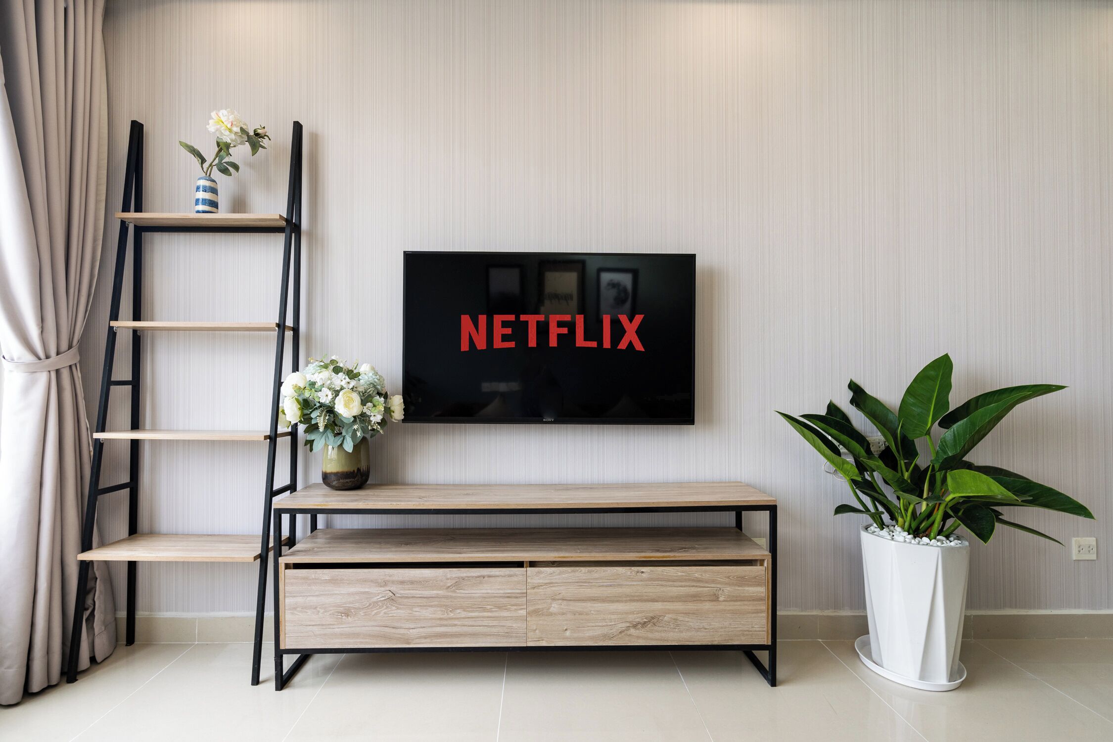 A modern living room with a TV mounted on the wall displaying the Netflix logo, a wooden bench, a potted plant, and a ladder against the wall.