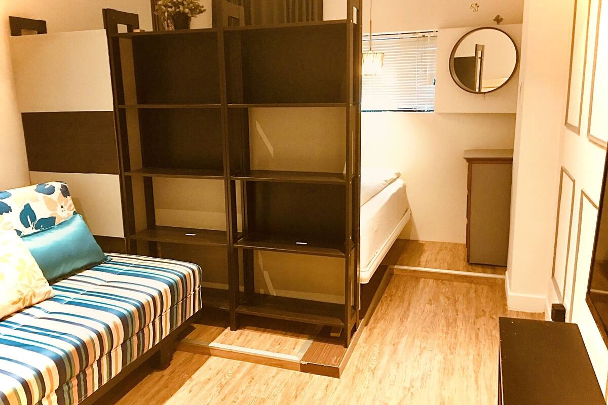 A compact bedroom with a bed, a small bedside table, a wardrobe, a mirror, and a window with blinds.