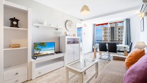 A modern living room with a flat-screen TV, white shelving, a glass coffee table, and a view of a cityscape through large windows. - Don Miguel II 4-F (Benidorm)