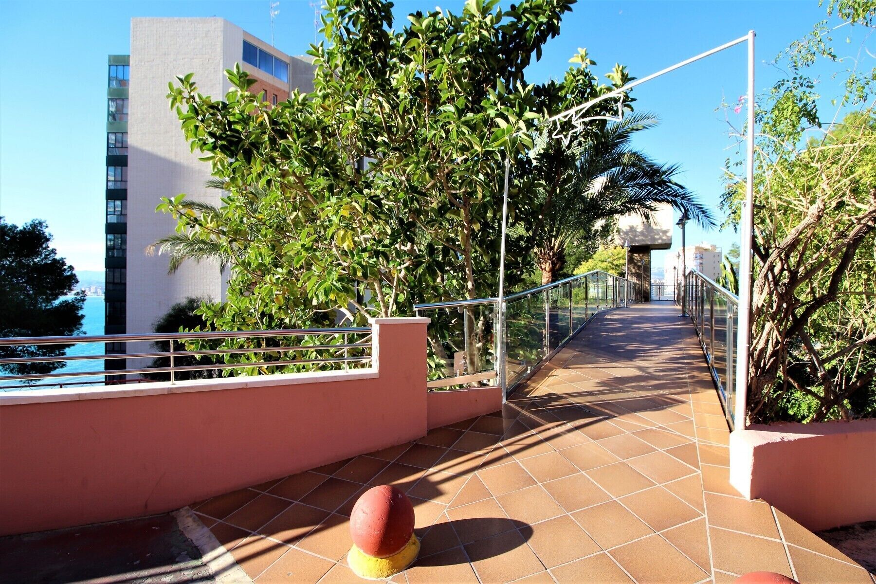 A pathway with a glass railing, surrounded by greenery and a building in the background.
