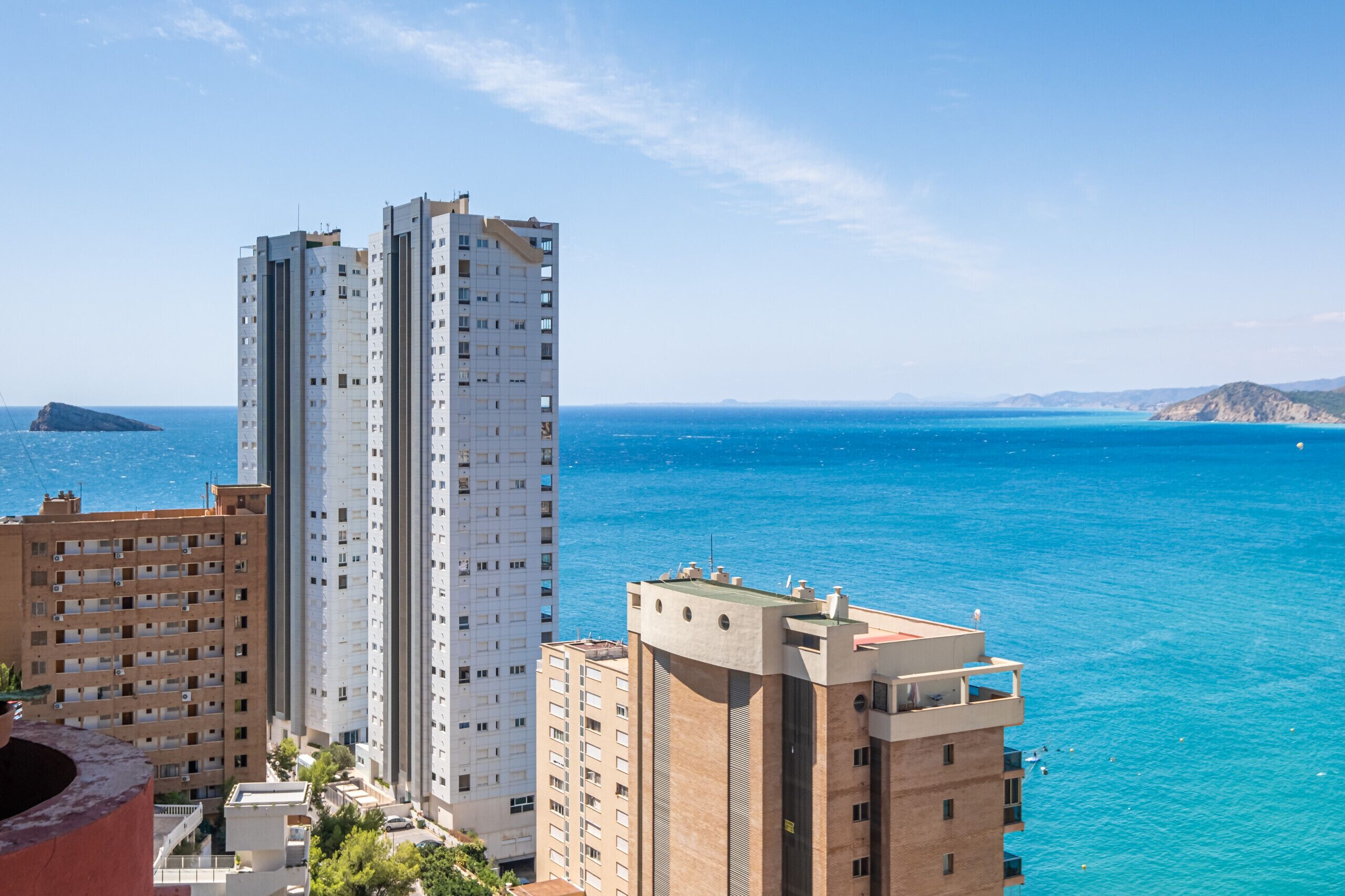 A coastal cityscape with high-rise buildings, including a prominent white tower and a brown building with balconies, overlooking a clear blue sea.