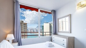 A hotel room with a bed, a nightstand, a lamp, a dresser, a window with curtains, and a view of a cityscape. - Don Miguel II 4-F (Benidorm)