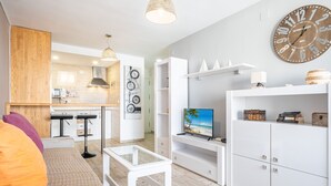 A modern living room with a sofa, coffee table, and TV. There is a kitchen in the background with a microwave and a hanging light fixture. - Don Miguel II 4-F (Benidorm)
