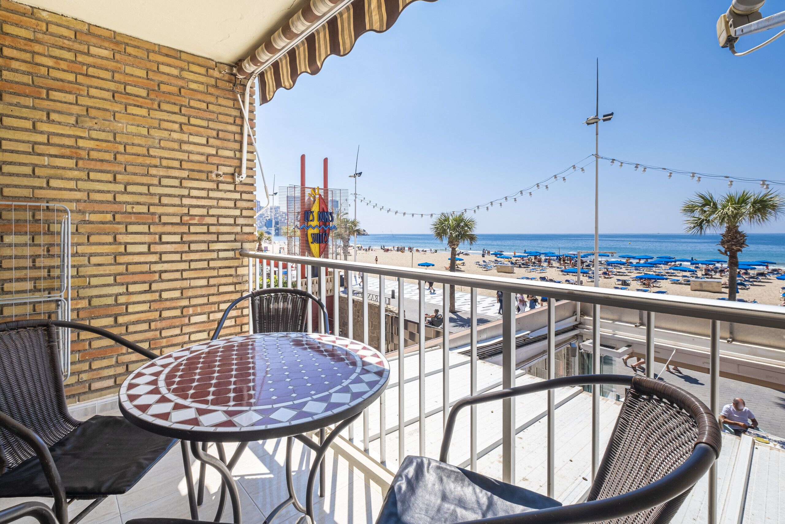 A balcony with wicker chairs and a tiled table overlooking a beach with umbrellas and palm trees.