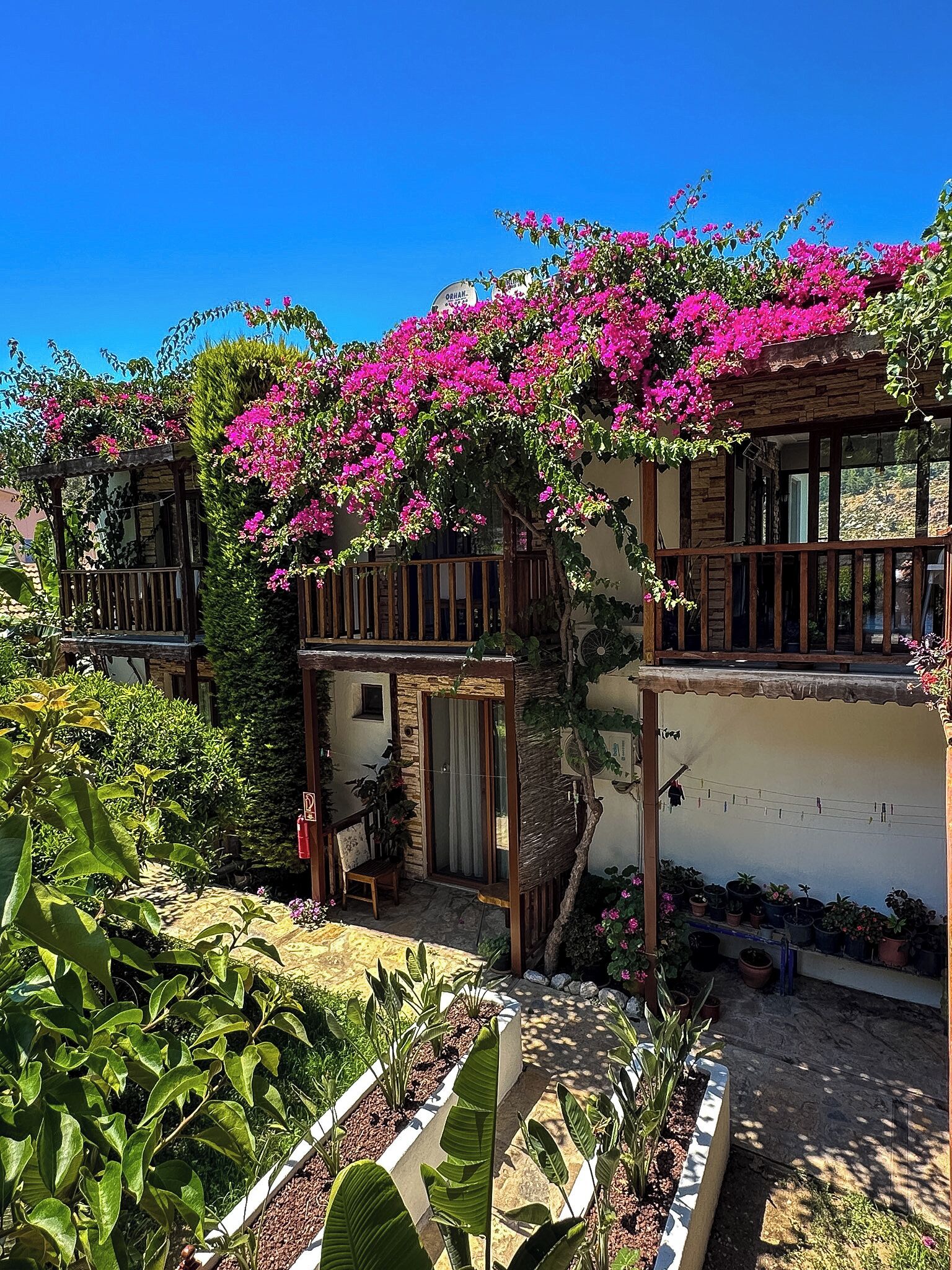 Un edificio de dos plantas con un balcón, un patio cubierto y un jardín con diversas plantas.