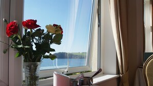 Uma mesa com um vaso de rosas, um jarro branco e uma pequena tigela, perto de uma janela com vista para o mar.