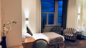 Quarto de hotel com uma cama, duas poltronas listradas, uma pequena mesa com abajur, um vaso com flores e uma janela com cortinas.