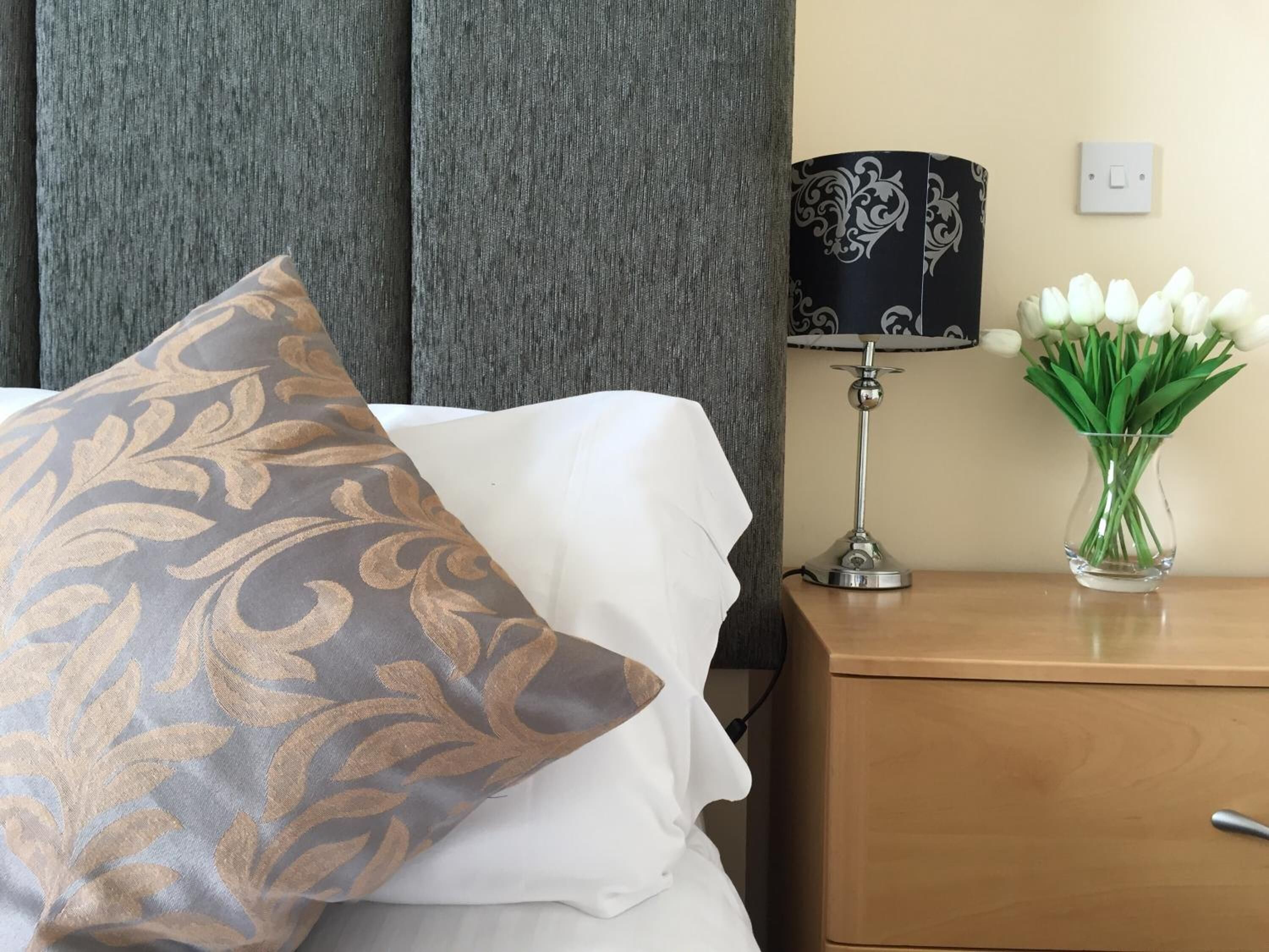 Et hotelværelse med en seng, et natbord, en lampe og en vase med blomster.