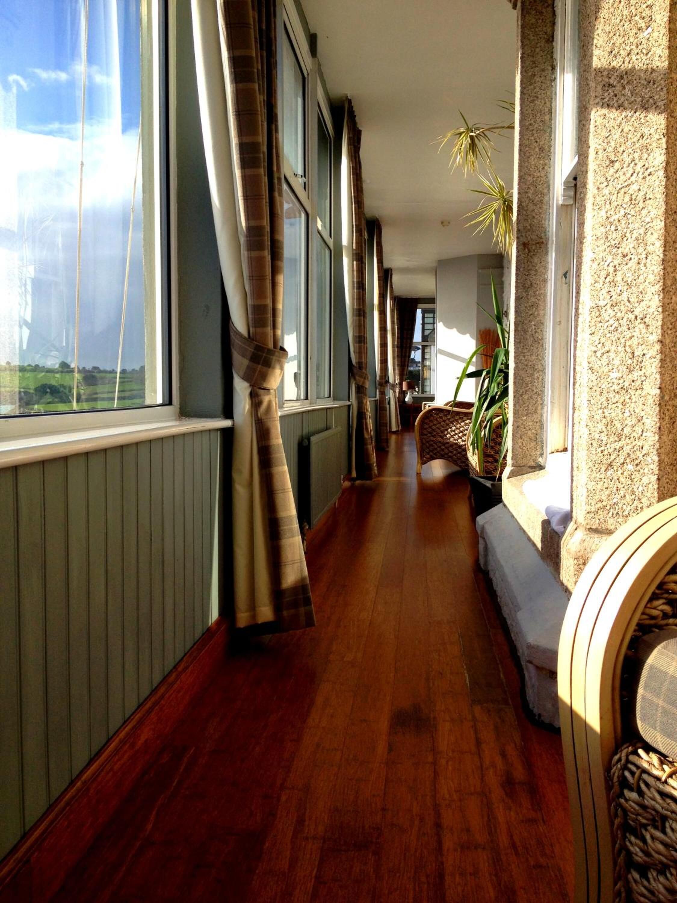 A corridor with wooden flooring, large windows with curtains, a bench, and a plant.