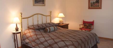 A neatly made bed with a patterned bedspread, two bedside lamps, a small bedside table with a lamp, a chair with a cushion, and two framed pictures on the wall.