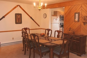 Un comedor con mesa y sillas de madera, una lámpara de candelabro, un reloj y una cocina al fondo.