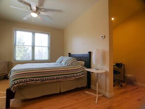 A bedroom with a bed, a standing fan, a small table, and a window.