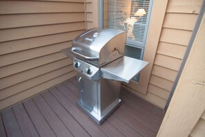 Een barbecue van roestvrij staal met een bijzettafeltje, geplaatst op een houten terras voor een huis met beige gevelbekleding.