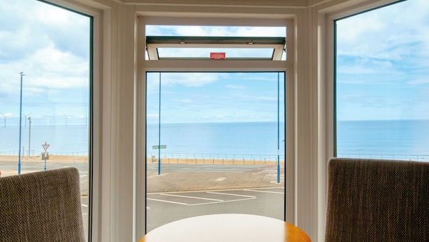 A room with a large window overlooking a coastal road and the sea, featuring two chairs and a small table.