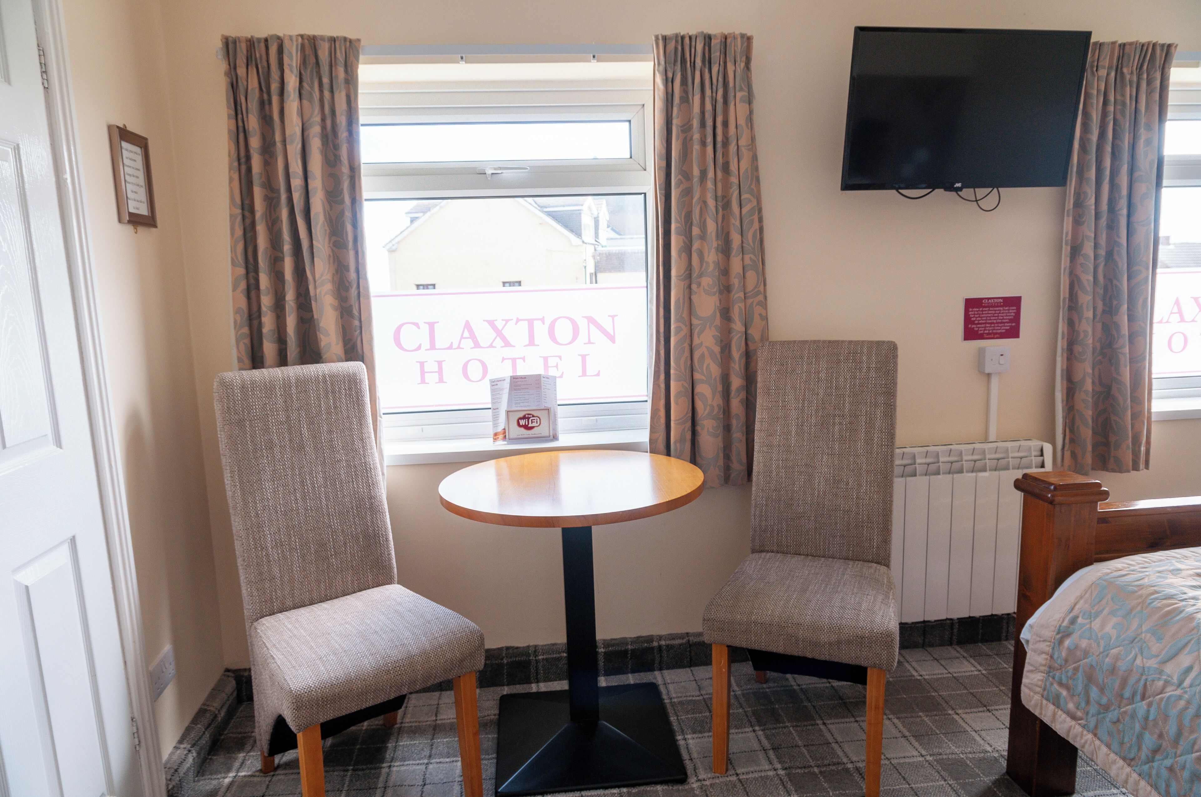 A hotel room with a plaid floor, two upholstered chairs, a small round table, a bed, a TV mounted on the wall, and a window with curtains.