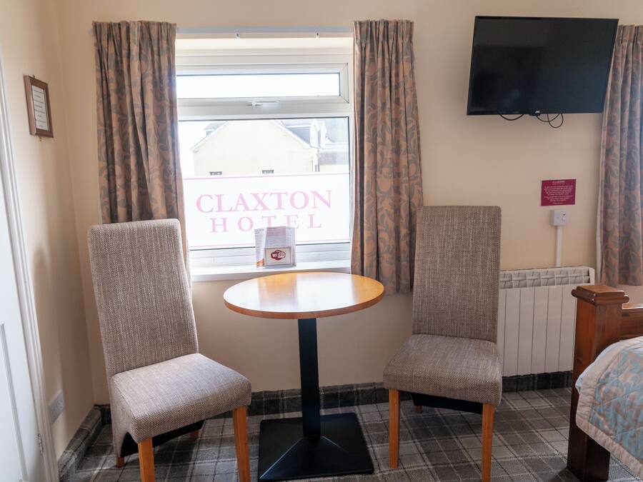 A hotel room with a plaid floor, two upholstered chairs, a small round table, a bed, a TV mounted on the wall, and a window with curtains.