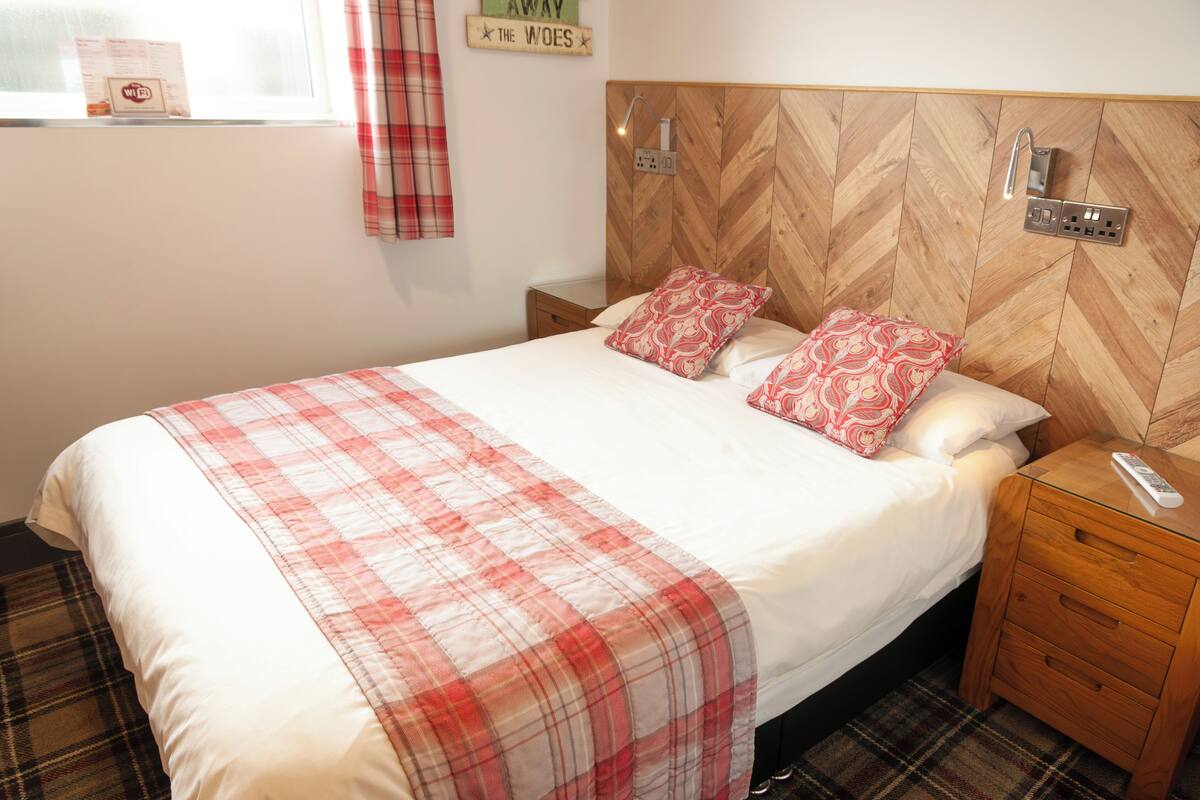 Un lit bien fait, recouvert d’une couverture à carreaux, avec deux oreillers à motifs rouges et blancs, une tête de lit en bois intégrant une lampe et une table de chevet avec une télécommande.
