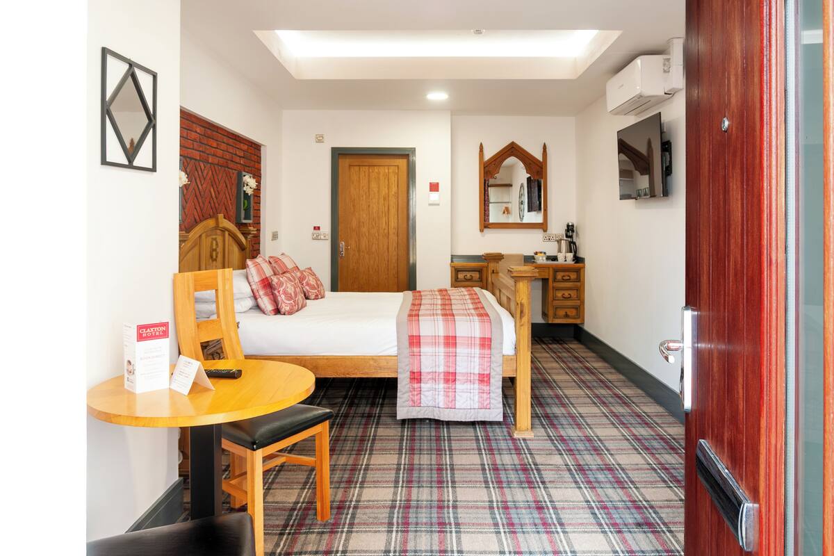 Une chambre d’hôtel avec un lit, un bureau en bois, une chaise, une petite table, un miroir et un sol à motifs écossais.