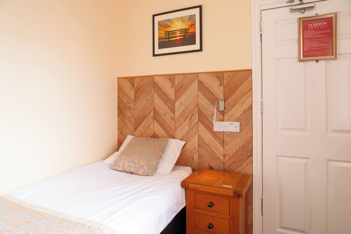 Un lit bien fait, avec une tête de lit en bois, une table de chevet dotée d’un tiroir, une prise électrique fixée au mur et un tableau représentant un coucher de soleil sur un banc.