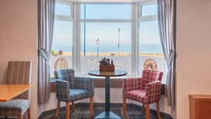 A corner room with a bay window overlooking the sea, featuring two plaid chairs, a small round table, and a wooden bench.