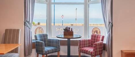 A corner room with a bay window overlooking the sea, featuring two plaid chairs, a small round table, and a wooden bench.