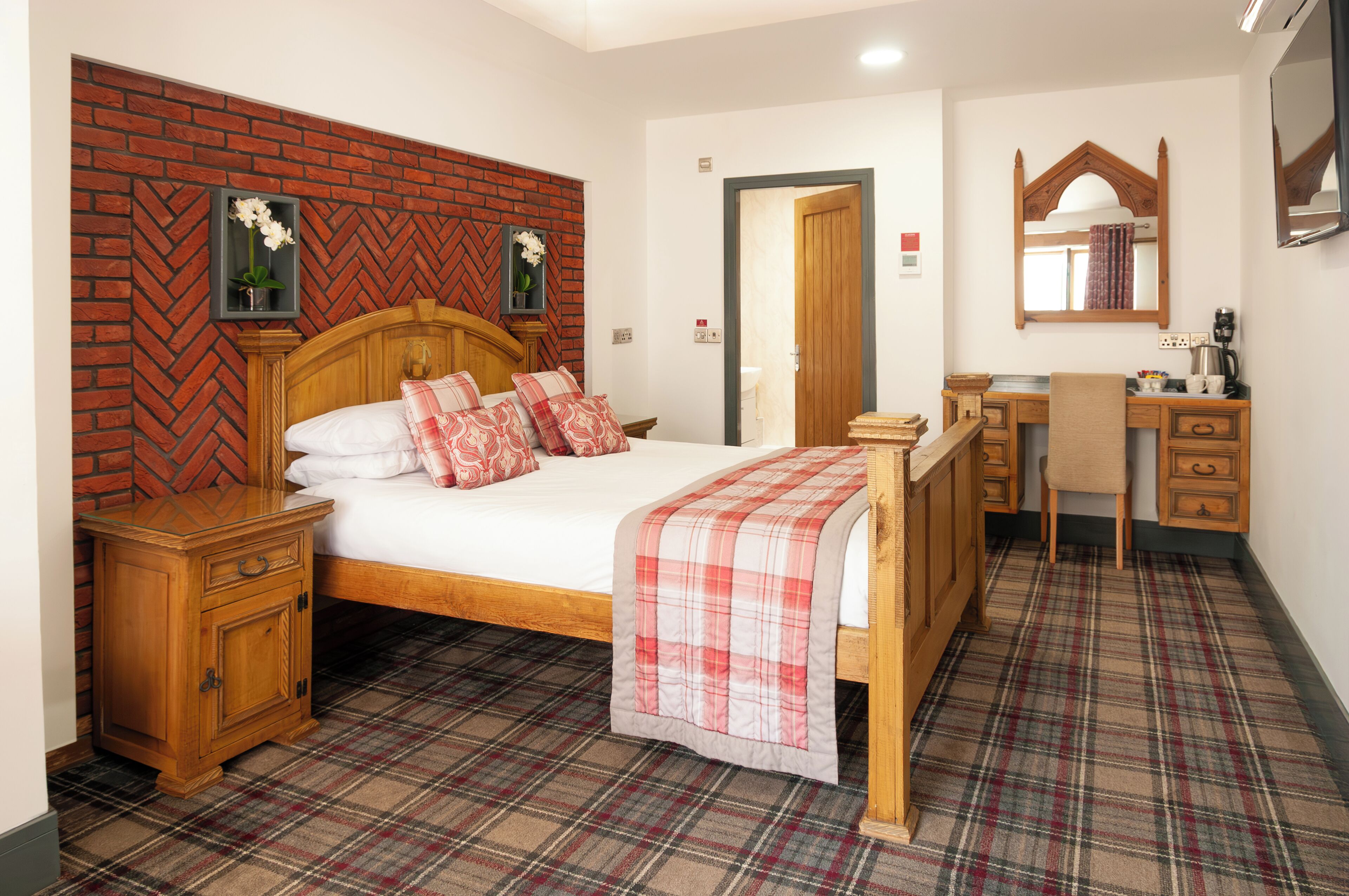 Un dormitorio con pared de ladrillo en patrón de espiga, una cama de madera con ropa de cama escocesa, una mesita de noche, un escritorio con una silla y un espejo.