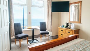 A hotel room with a bed, a desk with two chairs, a dresser, a TV, and a window with a view of the sea.