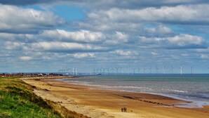 A beach with wind turbines in the distance, a sandy shore, and a cloudy sky.