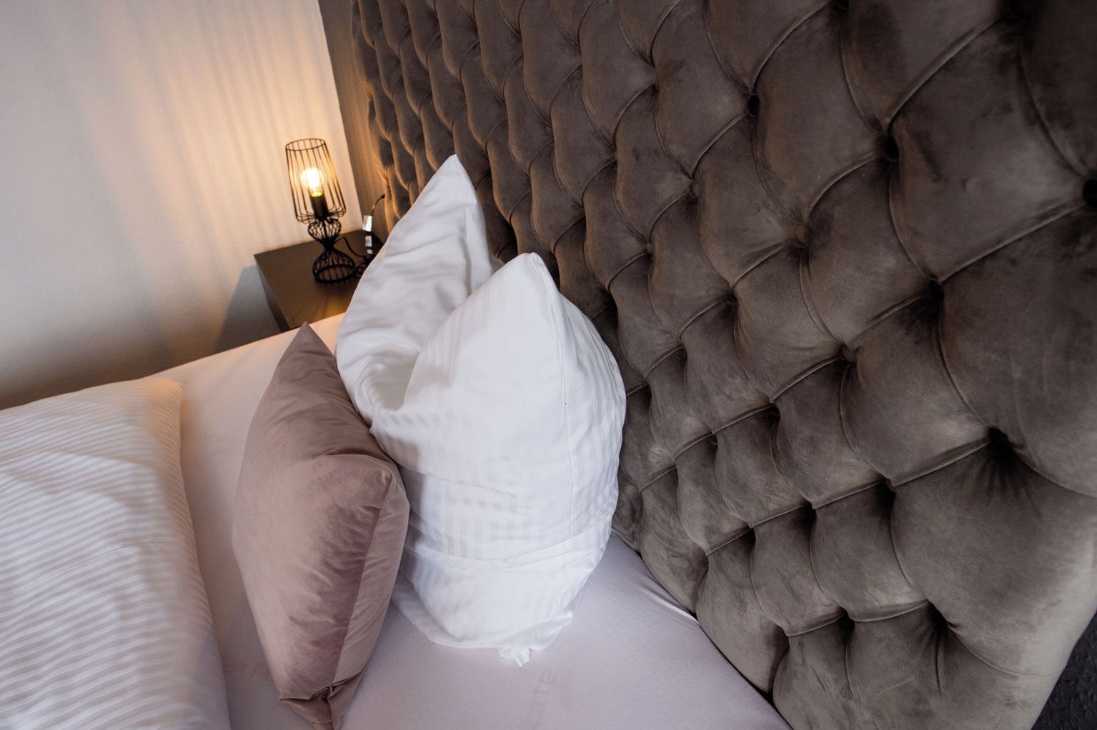 Ein Bett mit geblattetem Kopfteil, weißen und beige farbenen Kissen und einem Nachttisch mit Lampe.