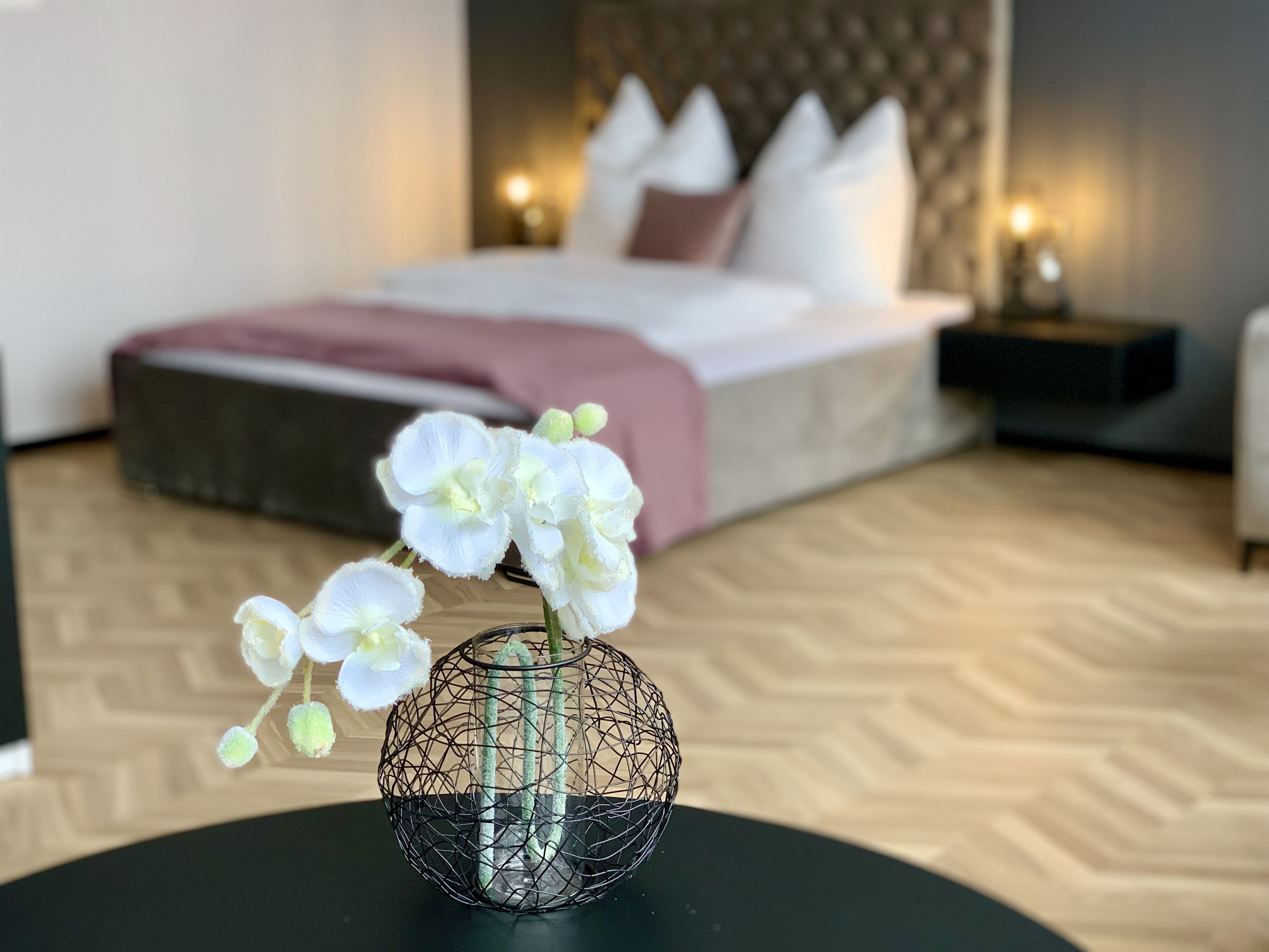A bedroom with a bed, bedside tables, and a vase with white flowers on a round table.