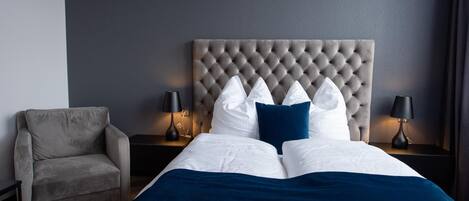 Una camera da letto con testiera trapuntata, un letto con biancheria bianca e azzurra, una poltrona grigia e due lampade da comodino.