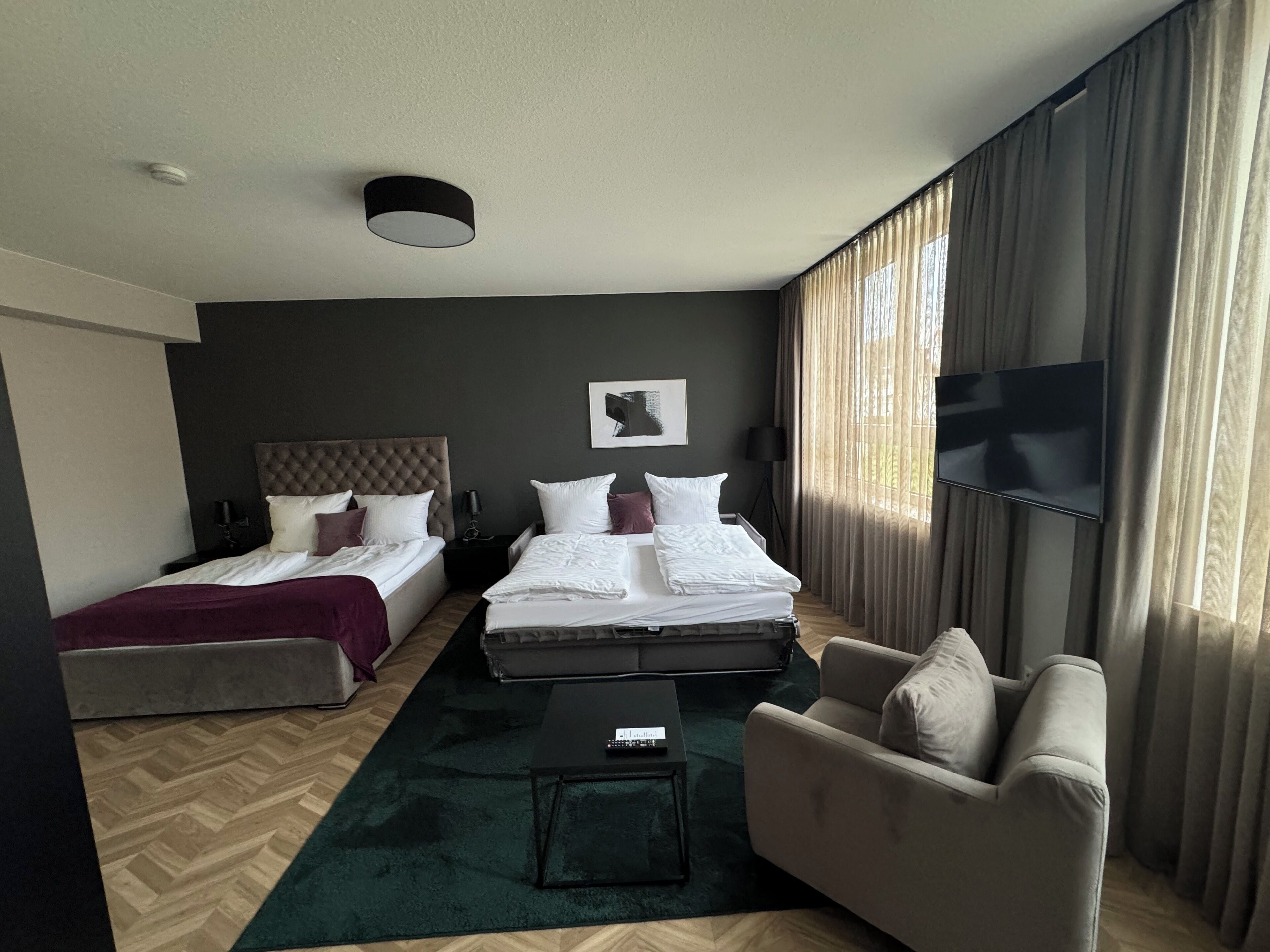 Une chambre d’hôtel avec deux lits, un canapé, une table basse, une télévision et une grande fenêtre avec des rideaux.