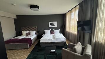 Habitació d'hotel amb dos llits, un sofà, una taula de cafè, una televisió i una finestra gran amb cortines.