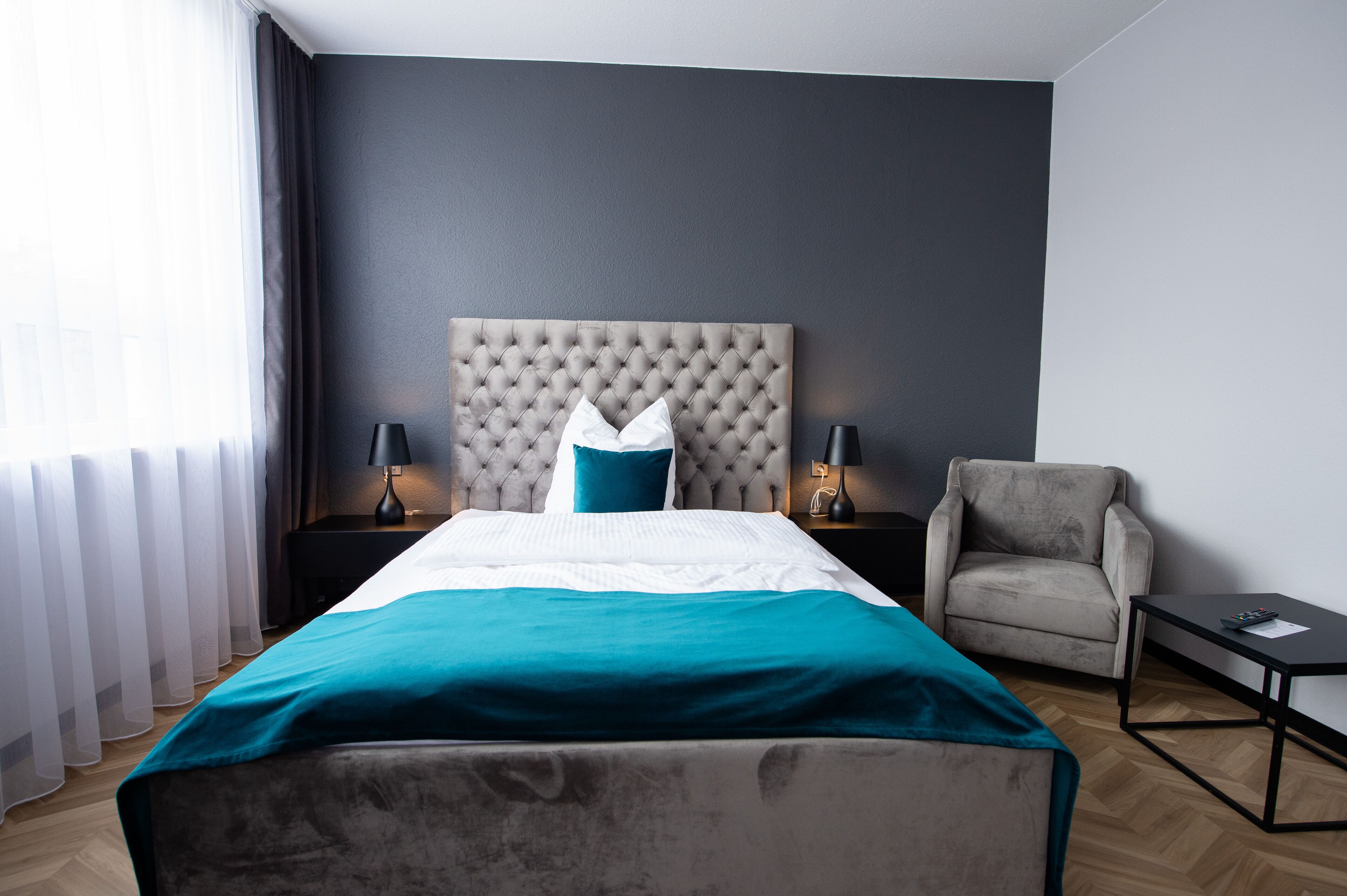 Una habitación de hotel moderna con cabecera capitonada, cama con ropa de cama color turquesa, un sillón gris, una mesita con un control remoto y una mesita de noche con lámparas.