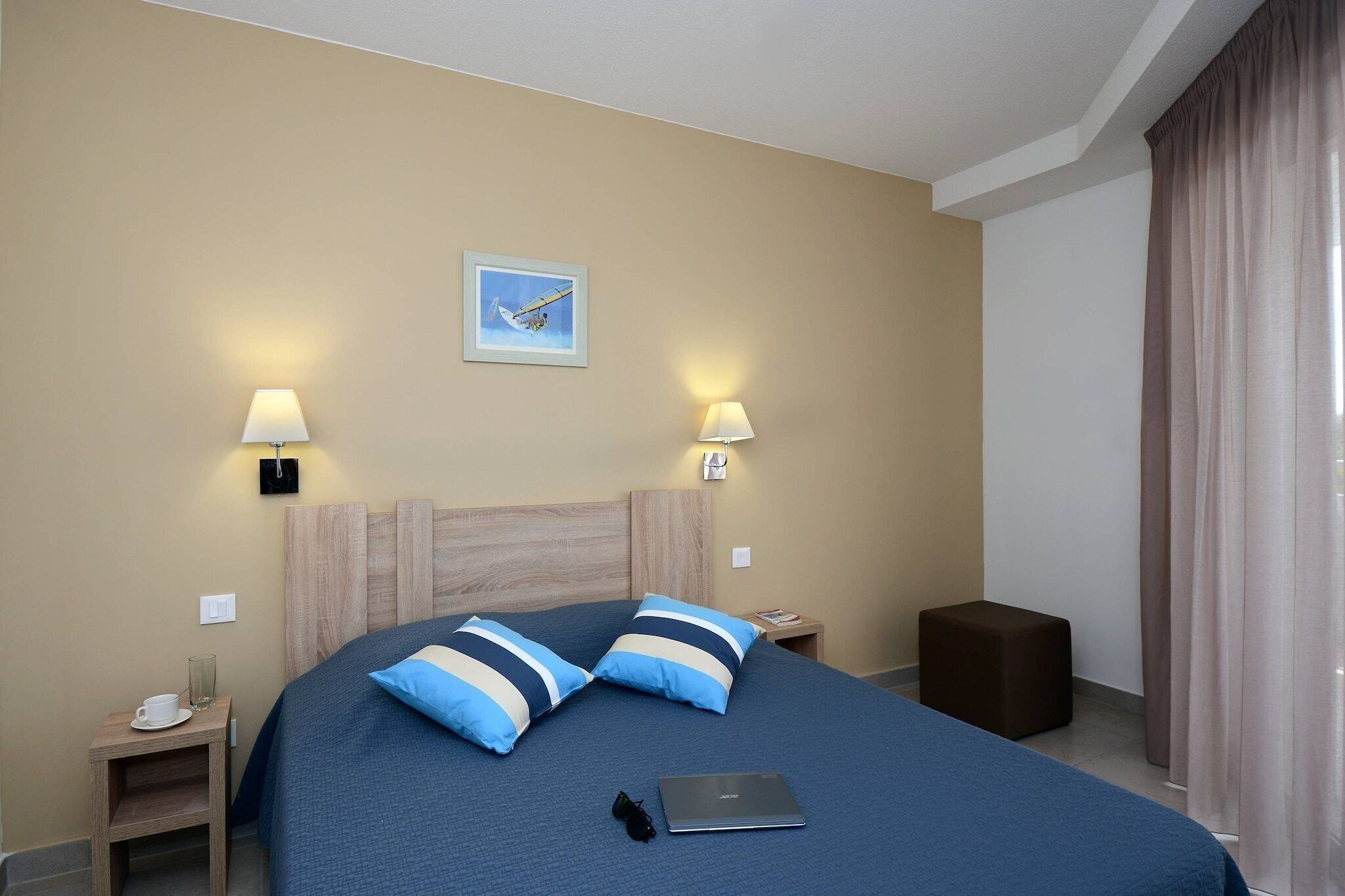 Habitación de hotel con cama, dos lámparas de noche, un cuadro en la pared, una laptop sobre la cama y un taburete de madera.
