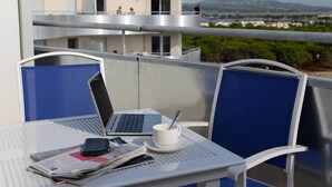En balkon med en bærbar computer, en kop og en ske på et bord, med udsigt over en bygning og et fjerntliggende landskab.